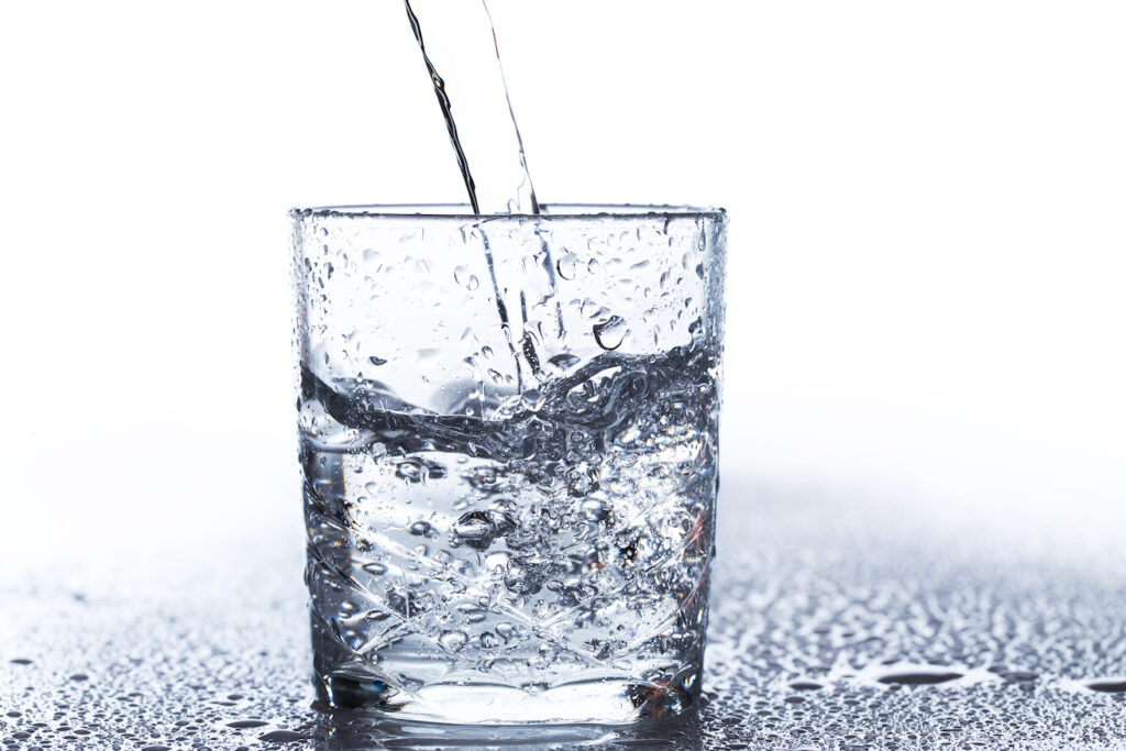 A stream of clear water is being poured into a glass, creating splashes and bubbles against a light background.