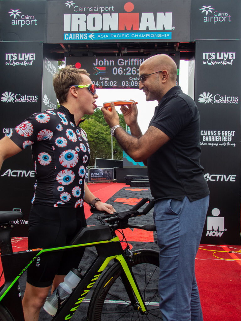 Two men interact at a cycling event, one in a colorful shirt offering a snack to the other beside a bike on a red carpet.