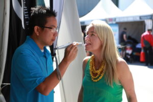 A woman with long blonde hair and a green top receives airbrush makeup from a person in a blue shirt at an outdoor event.