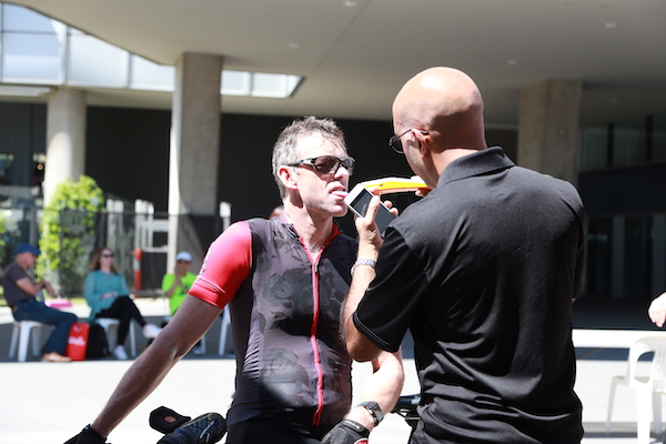 A cyclist in a tight-fitting jersey receives a drink from a coach outdoors, focused and preparing for competition.