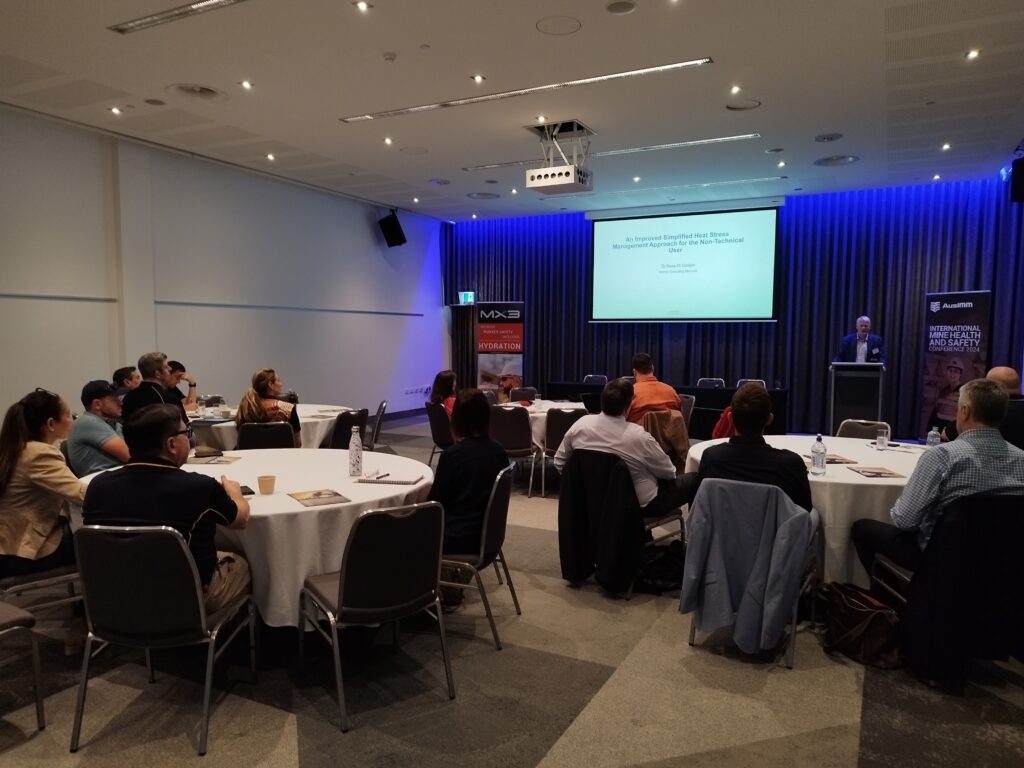 A conference room set up for a seminar, with attendees seated at round tables and a presenter speaking to an audience.