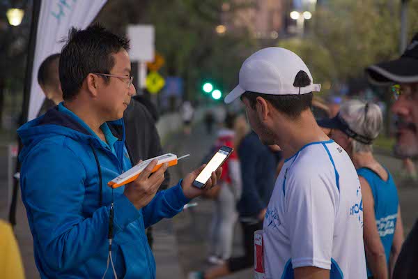 Two men converse outdoors; one holds a smartphone while the other listens, both engaged in discussion amidst a bustling background.