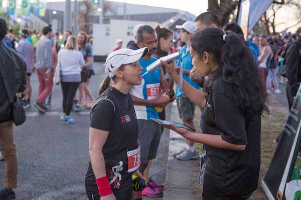 A runner in a black shirt and cap engages with a volunteer, while others prepare for a race in the background.