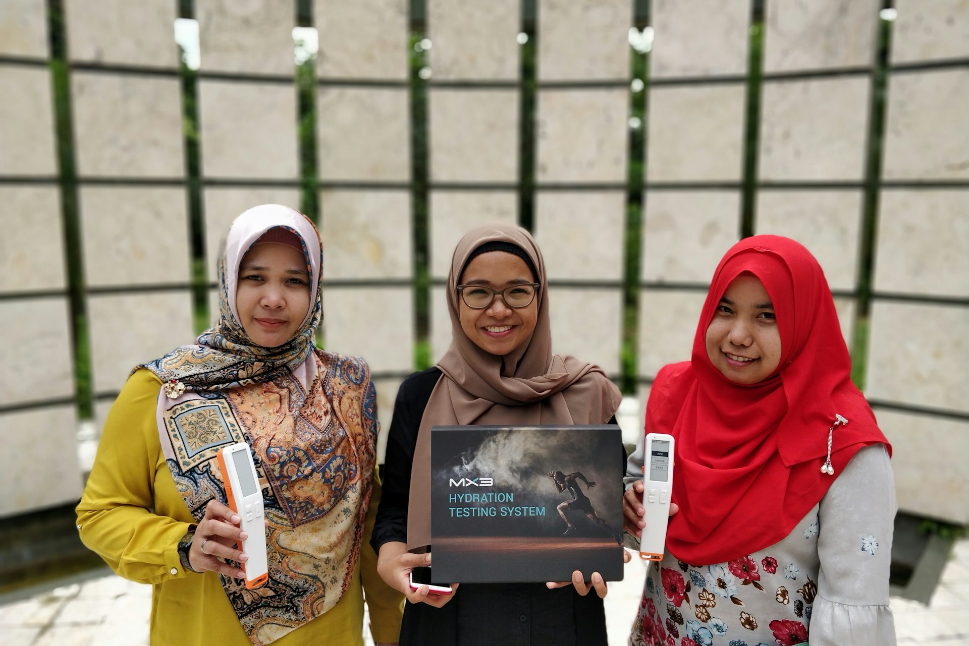Three women in colorful hijabs stand together, holding prizes and a plaque in front of a modern, textured background.