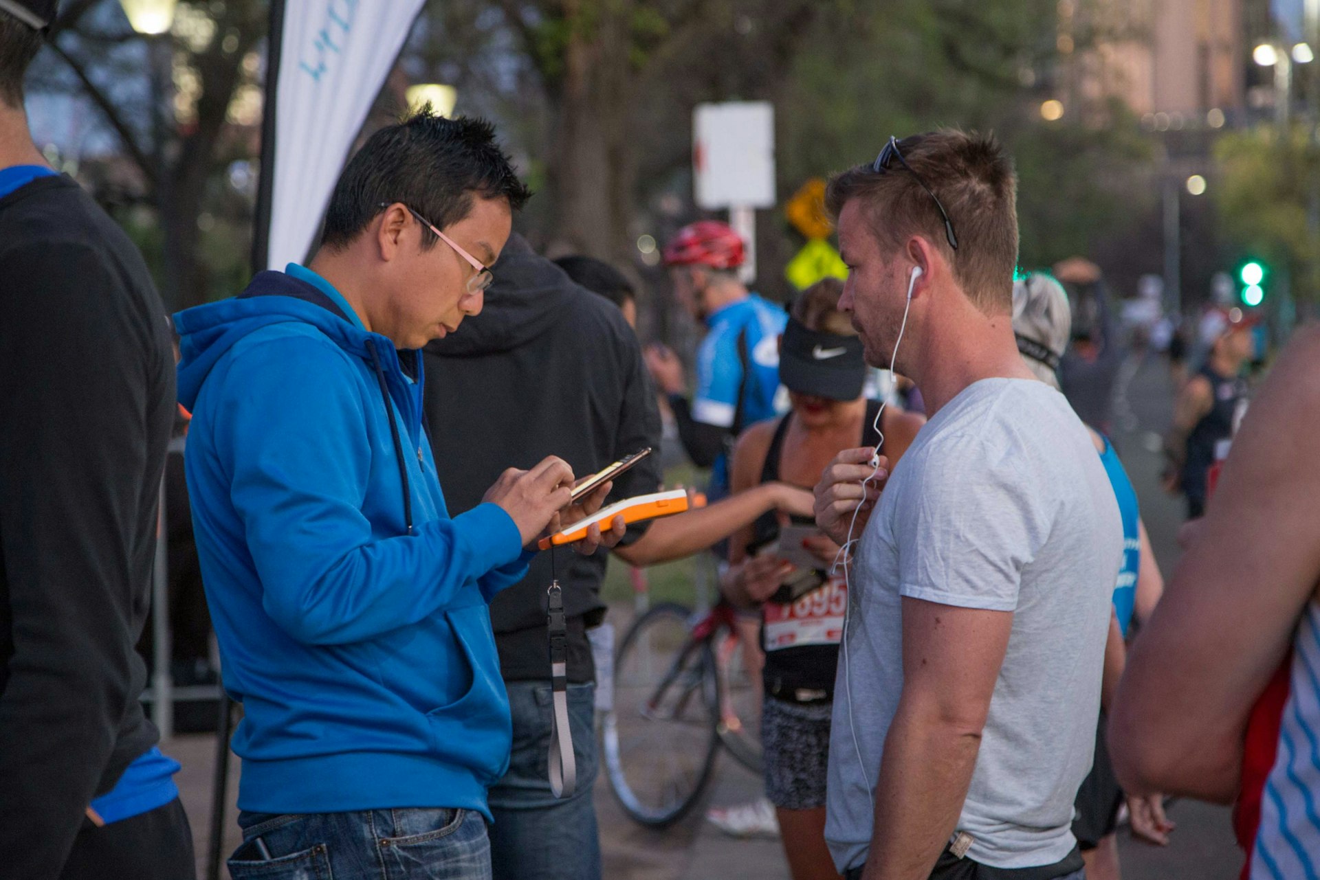 Two men converse at an outdoor event; one takes notes while the other holds a food item, with cyclists and bystanders in the background.
