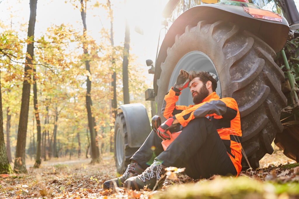 A forest worker in bright orange safety gear takes a break sitting against a large tractor tire amidst autumn foliage.