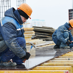 Two construction workers in orange helmets are laying wooden boards on a building site, with stacks of materials in the background.