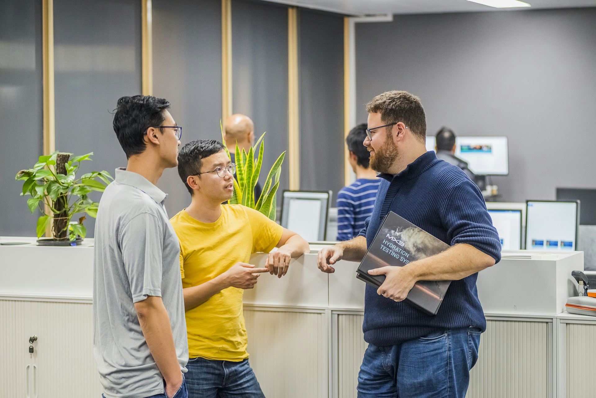 Three men engage in a discussion at a modern office, surrounded by plants and a welcoming atmosphere. One holds a tablet.