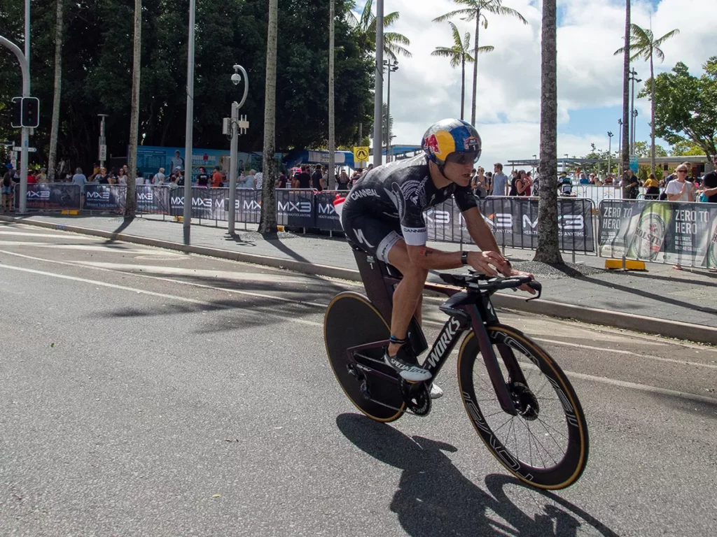 A cyclist in a black and yellow outfit speeds past spectators under palm trees during a race on a sunny day.