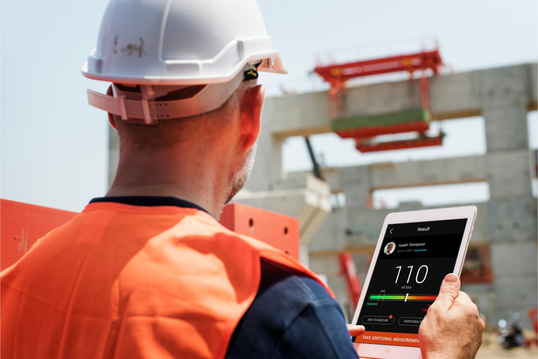 A construction worker in a hard hat and safety vest monitors equipment metrics on a tablet at a building site.